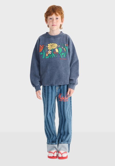 Enfant aux cheveux roux portant un sweat-shirt bleu à fleurs, un jean rayé avec un texte rouge, et des sandales rouges avec des chaussettes blanches, debout devant un fond uni.