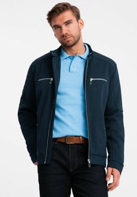 Veste zippée navy avec deux poches sur la poitrine, portée sur un polo bleu clair. Texture lisse, design ajusté, associée à un jean en denim foncé.