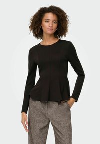 Kvindelig med krøllet hår iført en tætsiddende sort langærmet top og grå strukturerede bukser, stående med den ene hånd i lommen mod en hvid baggrund.