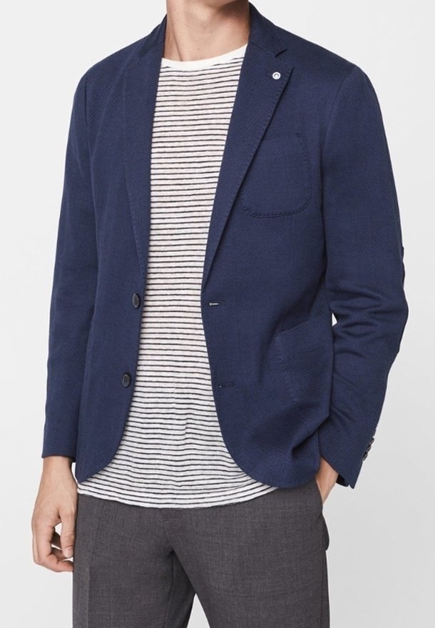 Homme portant un blazer bleu marine sur une chemise blanche rayée noire avec un pantalon gris, debout devant un fond blanc uni.
