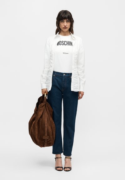 Γυναίκα που φορά λευκό πουκάμισο Moschino Milano, λευκή μπλούζα με βολάν, μπλε τζιν, μαύρα πέδιλα με ανοιχτά δάχτυλα, κρατώντας μεγάλη καφέ σουέντ τσάντα σε λευκό φόντο.