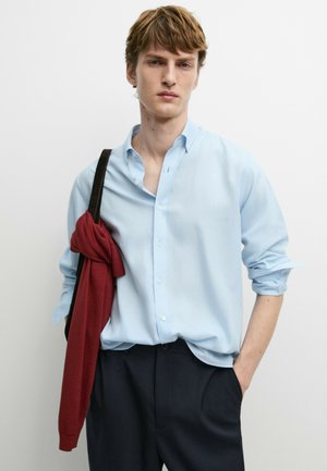 Giovane uomo che indossa una camicia azzurro chiaro con maniche arrotolate e pantaloni neri, porta una borsa nera con un maglione rosso appoggiato sopra.