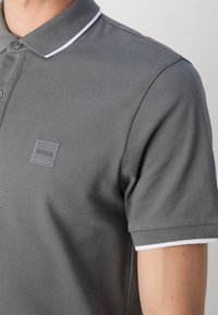 Gros plan d'une personne portant un polo gris avec une bordure blanche au col et aux manches, orné du logo en relief « BOSS » sur la poitrine.