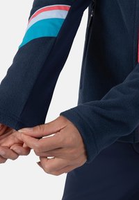 Giacca in pile color navy con zip frontale e strisce colorate rosa e turchese sulle maniche. Presenta un polsino aderente e una consistenza morbida.