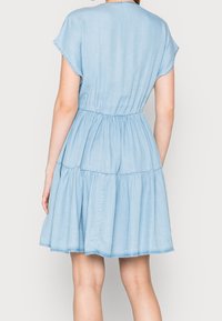 Vaaleansininen chambray-mekko lyhyillä hihoilla, vyötäröltä pussitetulla ja kerroksellisella hameella. Takana on pyöreä pääntie ja pehmeä kangas.