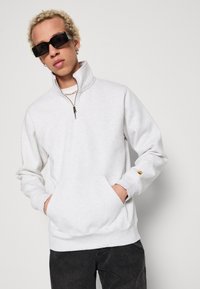 Lys grå sweatshirt med halv lynlås krave, kængurulommer og ribbede manchetter. Har en lille logo detalje på ærmet.