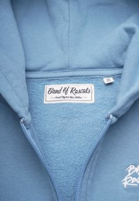 Ljusblå zip-up hoodie tillverkad av mjuk tyg. Har en fleecefoder invändigt och en lapp som säger "Band of Rascals."