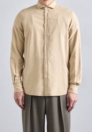 Homme portant une chemise beige à manches longues boutonnée et un pantalon plissé gris, debout, les bras détendus le long du corps.