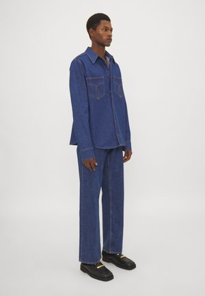 Denim shirt and pants set in a medium wash, featuring contrasting stitching, button-up front, chest pockets, and a relaxed fit. Black shoes with gold accent.