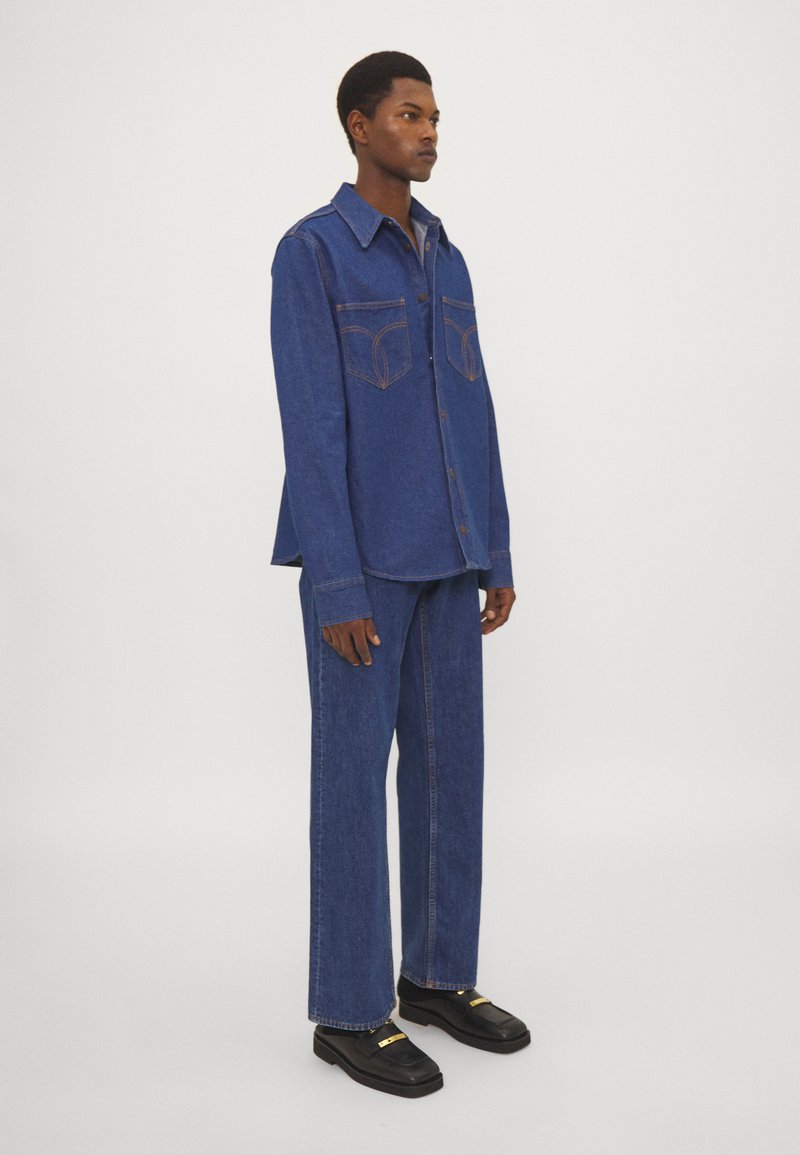 Set di camicia e pantaloni in denim di un lavaggio medio, con cuciture a contrasto, chiusura con bottoni, tasche sul petto e vestibilità rilassata. Scarpe nere con accenti dorati.