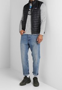 Gilet trapuntato nero sopra una maglietta grigia a maniche lunghe, abbinato a jeans azzurri con risvolti e scarpe da ginnastica verde scuro. Sfondo neutro.