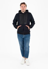 Felpa nera con zip e pannello frontale liscio, due tasche frontali, polsini e orlo a coste, abbinata a jeans blu chiaro e scarpe da ginnastica bianche.