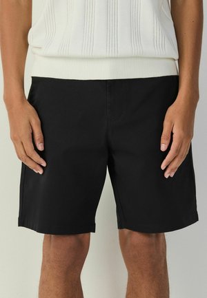 Man wearing black knee-length shorts and a white knit top, standing with hands relaxed at sides against a plain background.