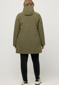 Veste imperméable vert olive avec capuche, coupe mi-longue, détails de couture, et leggings noirs associés à des baskets beiges.