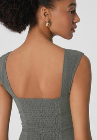 Robe grise texturée avec un décolleté carré et des bretelles larges. Portée par un mannequin montrant son dos, mettant en avant des accessoires pour les épaules et les oreilles.