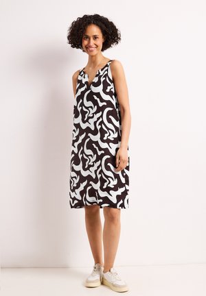 Kobieta o kręconych włosach uśmiechająca się, ubrana w czarno-białą bezrękawnikową sukienkę z abstrakcyjnym wzorem do kolan oraz białe trampki, stojąca na jednolitym tle.