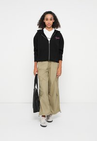 Hoodie zippé noir avec une poche avant, associé à un pantalon cargo baggy olive et des baskets claires. Tient un sac noir. Fond blanc.