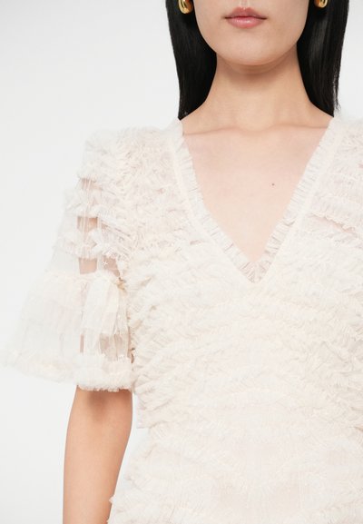 Kobieta ubrana w przezroczystą białą koronkową bluzkę z falbankowym wzorem, krótkimi rękawami i dekoltem w kształcie litery V, w zestawieniu ze złotymi kolczykami kołami.