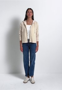 Felpa zip beige chiaro con cappuccio e cordini, abbinata a jeans a gamba larga blu medio e sneaker beige con dettagli marroni. Design semplice e informale.