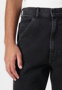 Black denim jeans with a faded finish, tapered fit, and front pockets. Features visible stitching and a button closure at the waistband.