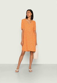 Vestido curto laranja com bolinhas brancas, decote em V, mangas bufantes e um corte descontraído. Combinado com sandálias brancas e uma mala de mão creme.