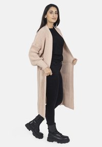 Cardigan lungo aperto in maglia rosa chiaro con texture a coste. Abbinato a una maglietta nera e pantaloni neri, e stivali da combattimento neri.