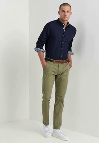 Camisa de botones azul marino con puños a rayas, pantalones verde oliva y zapatillas blancas. El cinturón marrón añade contraste al conjunto.