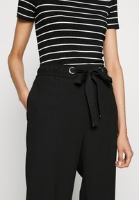 Femme portant un haut à manches courtes rayé noir et blanc, rentré dans un pantalon noir taille haute avec une ceinture nouée.