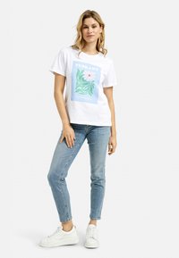 White t-shirt featuring a blue, green, and pink floral graphic that reads “TUSKANY FLOWER GARDEN.” Paired with light blue jeans and white sneakers.