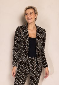 Schwarz-beiges Anzugset mit einem taillierten Blazer mit Reverskragen und schmal zulaufenden Hosen. Strukturierter Stoff mit geometrischem Muster.