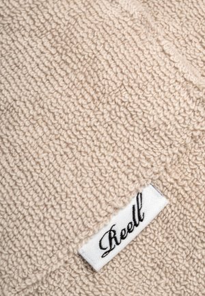 Beiger, strukturierter Stoff mit einer weichen, kuscheligen Optik und einem weißen Etikett, auf dem das Wort "Reell" in schwarzer Schreibschrift steht.