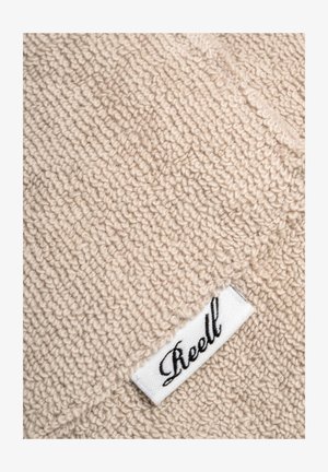 Beiger, strukturierter Stoff mit einer weichen, kuscheligen Optik und einem weißen Etikett, auf dem das Wort "Reell" in schwarzer Schreibschrift steht.