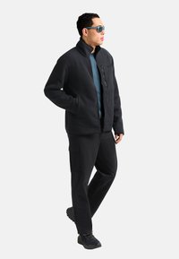 Giacca nera a costine con colletto alto, tasca sul petto con zip, sovrapposta a una maglia blu, abbinata a pantaloni cargo neri e scarponi da trekking.