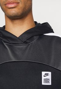 Sweat à capuche noir à la texture lisse, avec un patch logo Nike blanc distinct et une capuche prononcée. La partie supérieure est en matériau brillant.