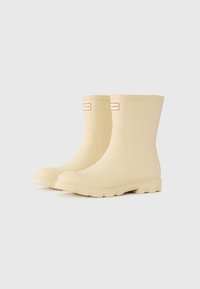 Bottes de pluie en caoutchouc imperméables beiges avec une finition lisse, des orteils ronds et des semelles texturées. Présente des étiquettes de marque rouges et blanches sur les côtés.