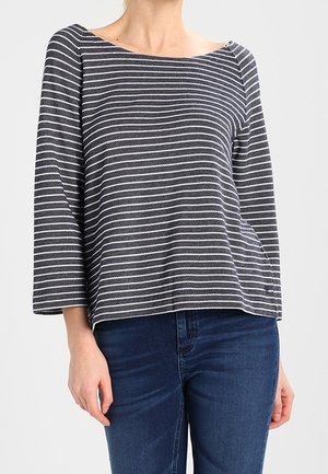 Kobieta ubrana w czarno-białą bluzkę w poziome paski z długim rękawem i dekoltem w łódkę oraz ciemnoniebieskie dżinsy.