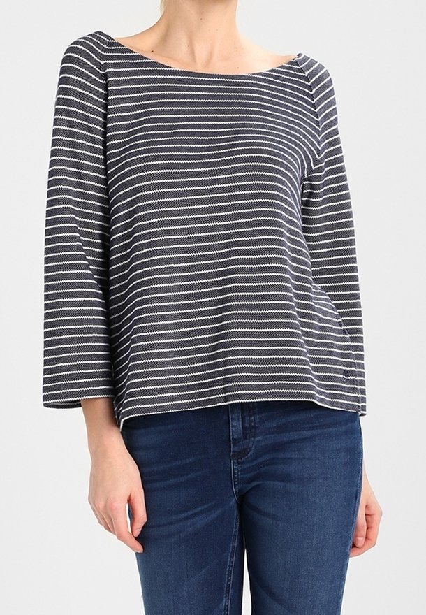 Femme portant un haut à manches longues à rayures horizontales noires et blanches avec un col bateau et un jean bleu foncé.