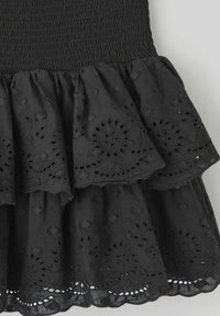 La robe noire présente un corsage en smock, un ourlet à volants en couches avec une broderie florale découpée, et une texture en coton douce.