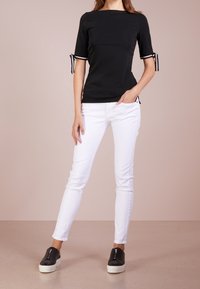 Černý přiléhavý top s krátkými rukávy zdobený bílými akcenty a mašlemi, v kombinaci s bílými úzkými kalhotami a černými nazouvacími botami.