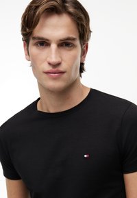 Junger Mann mit kurzen braunen Haaren, der ein schlichtes schwarzes T-Shirt mit einem kleinen rot-weiß-blauen Logo auf der Brust trägt.