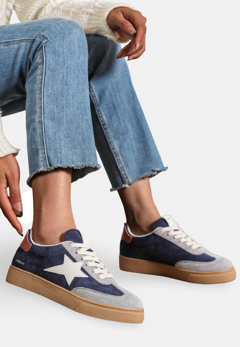 Baskets en daim navy et gris avec un détail en forme d'étoile blanche, semelle en gomme et lacets crème. Portées avec un jean bleu effiloché et un pull en tricot blanc.