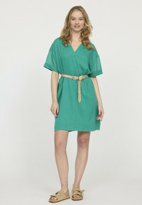 Robe courte verte avec un décolleté en V et des manches courtes, cintrée à la taille par une ceinture tressée beige. Tissu léger et coupe décontractée.