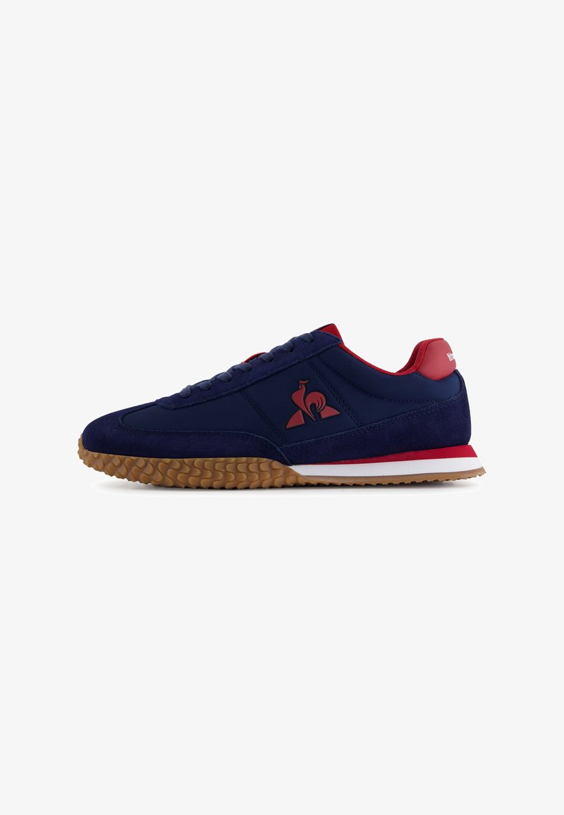Chaussures de sport bleu marine avec une tige en daim et en maille, accents rouges, semelle en gomme texturée et un logo sur le côté, présentant une silhouette classique.