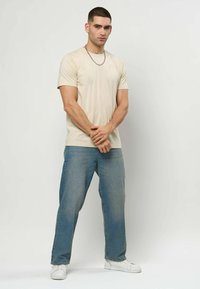 Beige bomull t-skjorte med korte ermer, kombinert med lyseblå jeans med rette ben og hvite joggesko. Har en liten logo på skjorten.