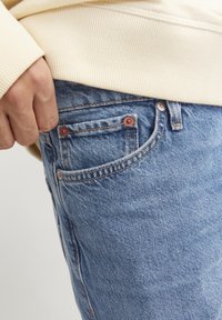 Svijetloplave trapera hlače sa bakrenim kopčama, detaljima na bočnim džepovima i vidljivim šavovima; ruka položena na bok, iznad duks u boji kreme.