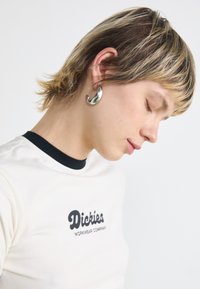 White T-shirt with black logo, high neckline, and a silver earring. Hair is short with blonde highlights. Clean, minimal background.
