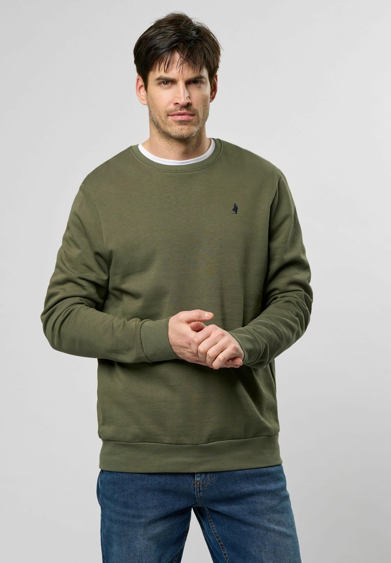 Zöld crewneck pulóver, pamutból készült, bordás mandzsettákkal és aljjal, a mellkason egy apró logóval. Fehér ing fölött viselve.
