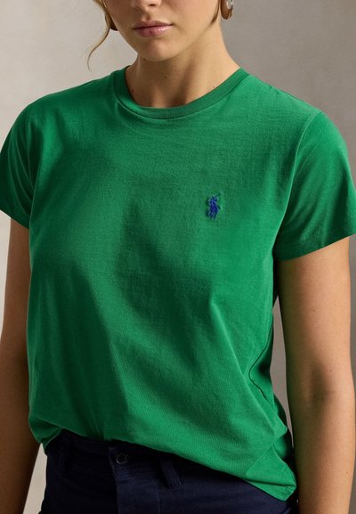 Mladá žena nosí zelené tričko s krátkym rukávom a malým modrým vyšívaným logom na ľavej strane hrudníka a tmavé nohavice.