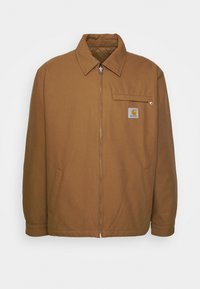 Chaqueta marrón con cremallera, hecha de tejido suave, con un cuello puntiagudo, bolsillos laterales y un bolsillo en el pecho con un parche de logo visible.