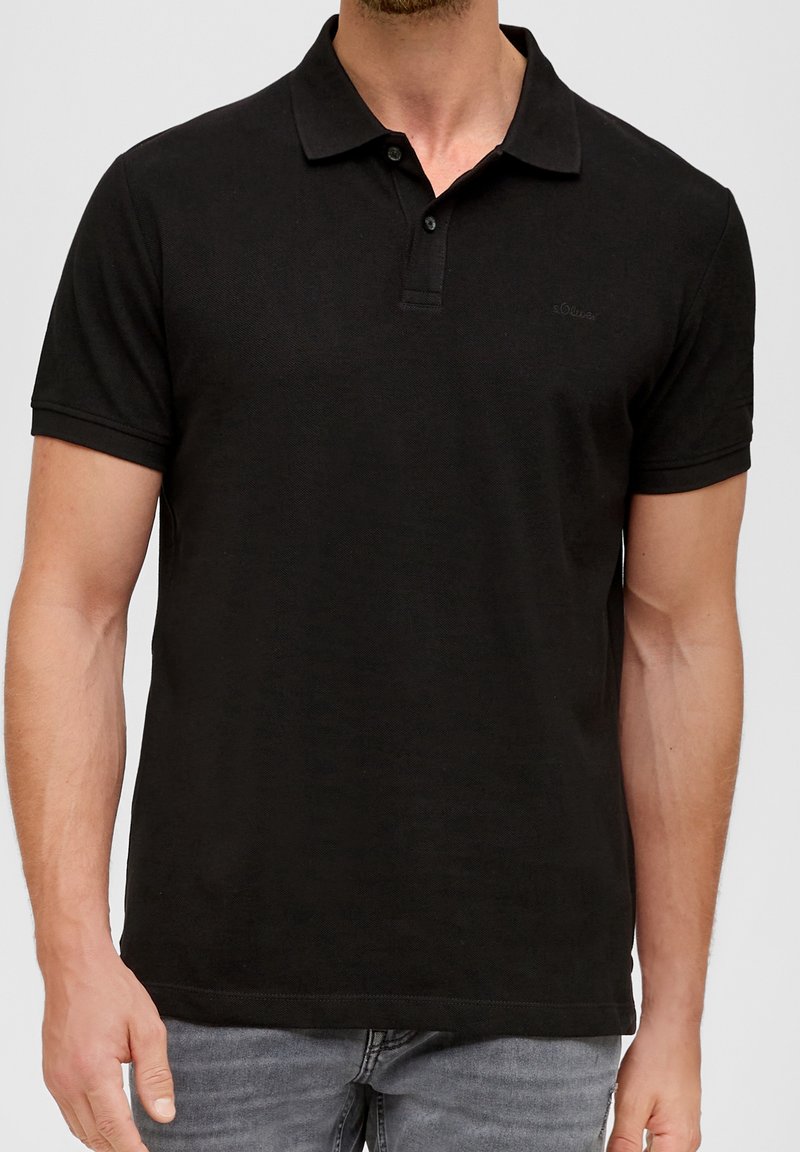 Homme portant un polo noir uni à manches courtes et un jean gris, debout devant un fond neutre.